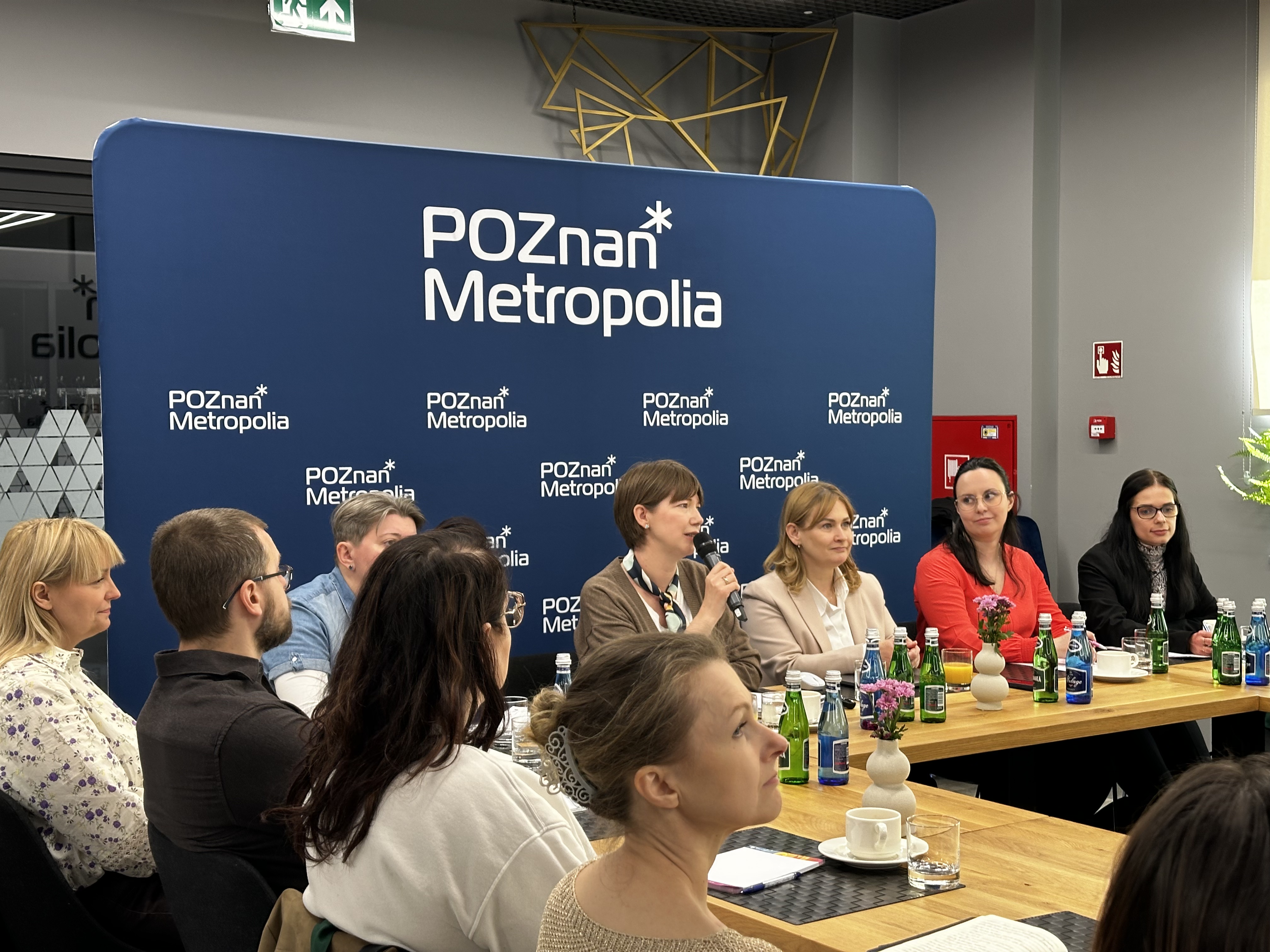 Zdjęcie przedstawia kilka osób siedzących obok siebie na tle ścianki. Na pierwszym planie kobieta, t