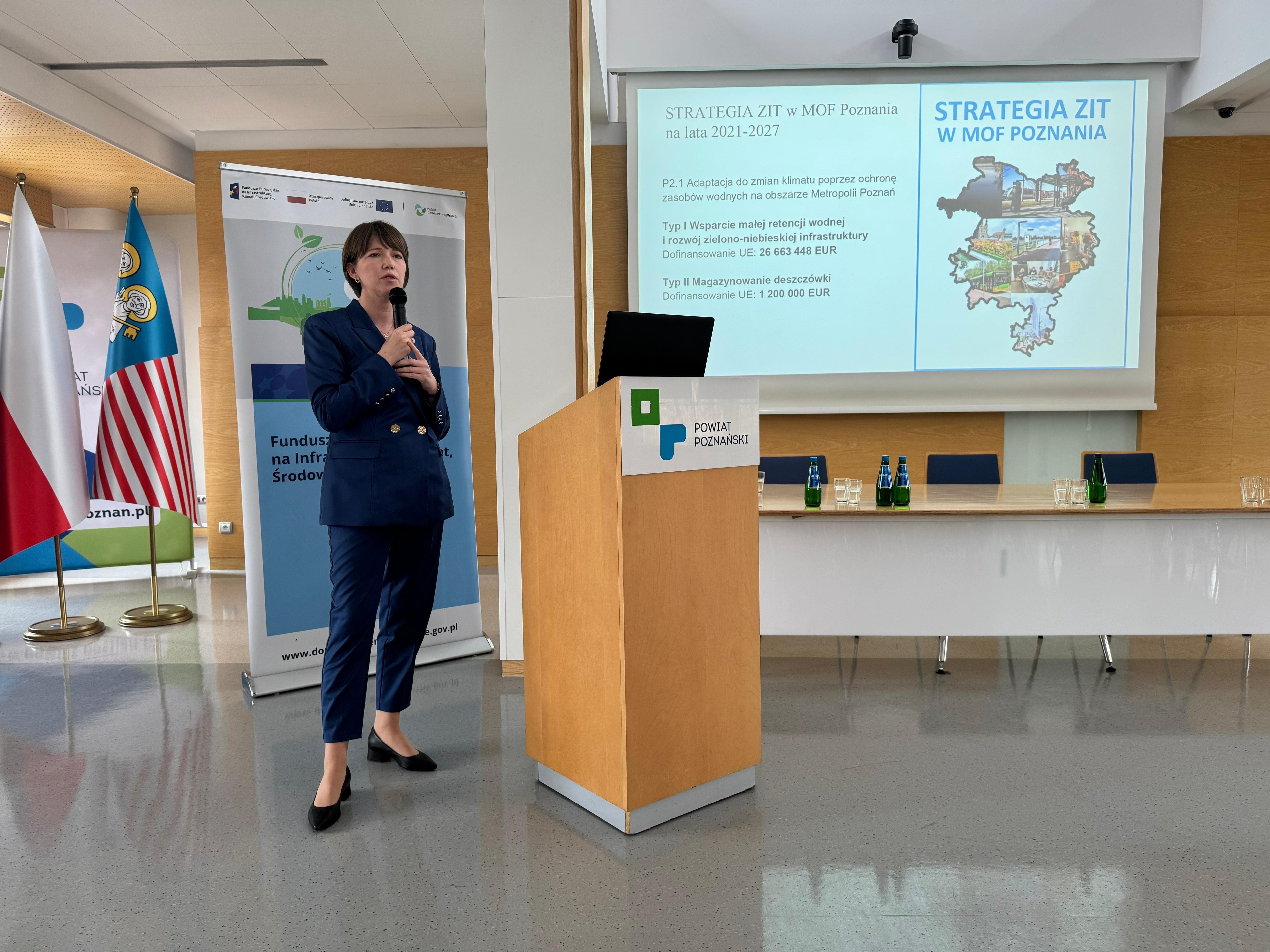 Zdjęcie przedstawia przemawiającą kobietę. W tle tle widać flagi oraz ekran.