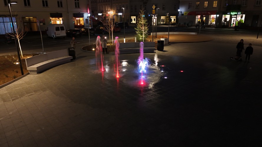 Widok Placu 20 Października w Śremie po zmroku z lotu ptaka. Plac oświetlony latarniami. W centralnej części widoczna podświetlona kolorami fontanna.