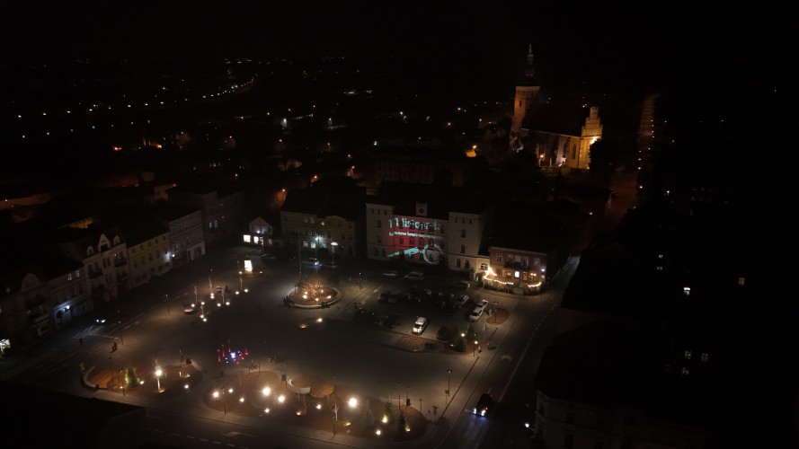 Widok Placu 20 Października w Śremie po zmroku z lotu ptaka. Plac oświetlony latarniami. W dolnej części widoczna podświetlona kolorami fontanna.