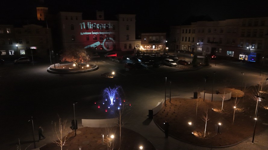 Widok Placu 20 Października w Śremie po zmroku z lotu ptaka. Plac oświetlony latarniami. W centralnej części widoczna podświetlona kolorami fontanna.