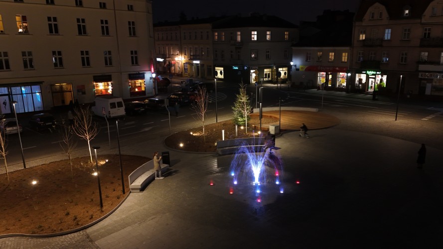 Widok Placu 20 Października w Śremie po zmroku z lotu ptaka. Plac oświetlony latarniami. W dolnej części widoczna podświetlona kolorami fontanna.