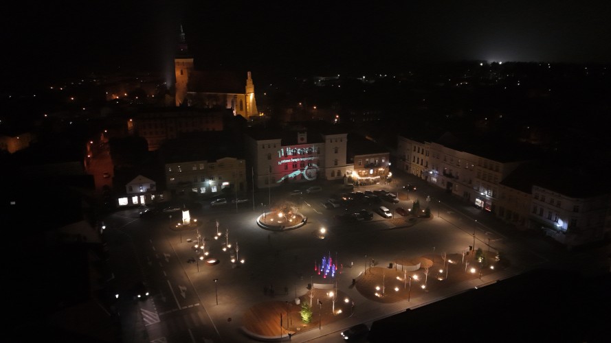 Widok Placu 20 Października w Śremie po zmroku z lotu ptaka. Plac oświetlony latarniami. W dolnej części widoczna podświetlona kolorami fontanna.