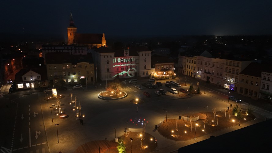 Widok Placu 20 Października w Śremie po zmroku z lotu ptaka. Plac oświetlony latarniami. W dolnej części widoczna podświetlona kolorami fontanna.