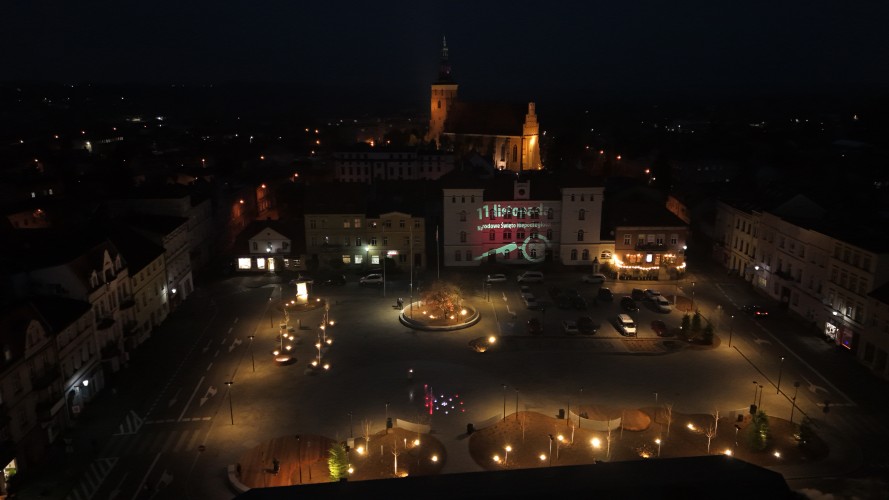 Widok Placu 20 Października w Śremie po zmroku z lotu ptaka. Plac oświetlony latarniami. W dolnej części widoczna podświetlona kolorami fontanna.