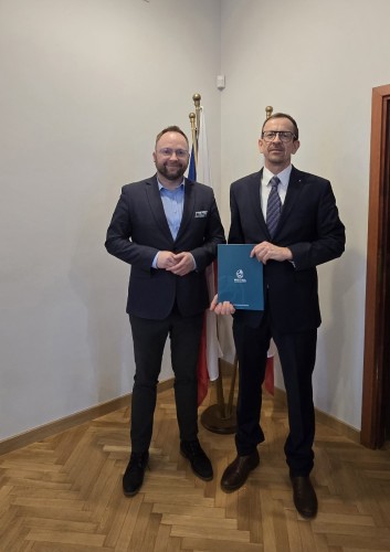 Fotografia pokazuje dwie osoby w garniturach prezentujące teczkę z podpisaną umową.