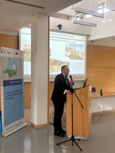 Zdjęcie przedstawia mężczyznę przemawiającego na tle ekranu.