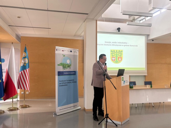 Zdjęcie przedstawia mężczyznę przemawiającego na tle flag oraz ekranu.