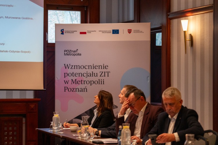 Zdjęcie przedstawia ludzi siedzących przy stole, a w tle widać ściankę z logotypami.