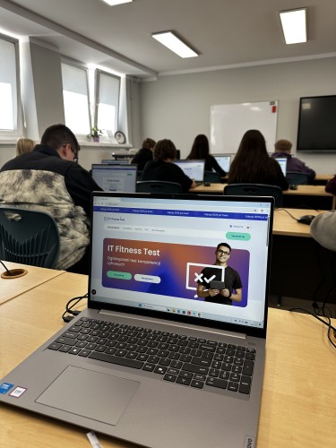Zdjęcie przedstawia grafikę IT Fitness Test na laptopie, na tle klasy.