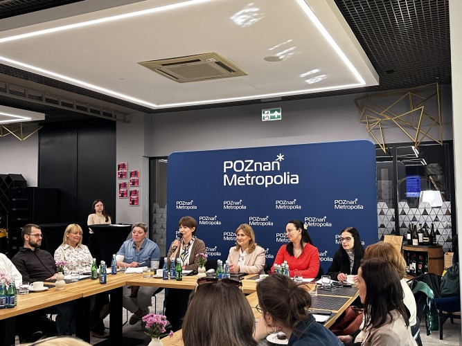Zdjęcie przedstawia kobietę przemawiającą z mikrofonem w ręce (zastępca dyrektora Metropolii Poznań). Wokół niej siedzą ludzie.