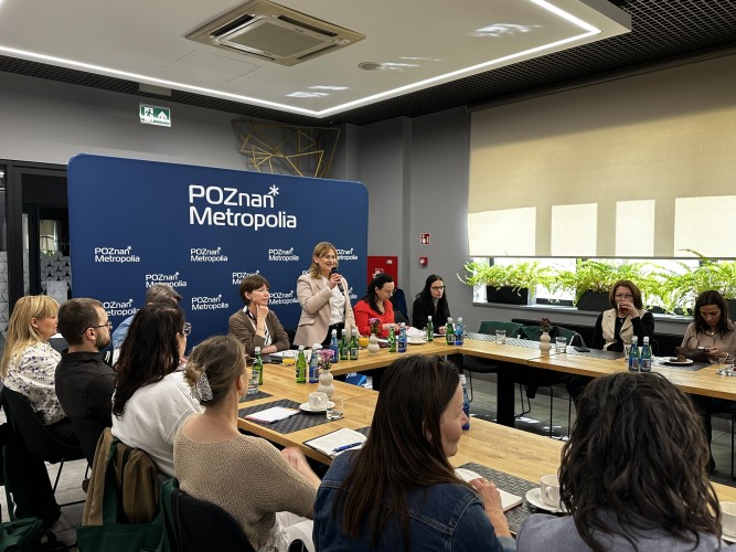 Zdjęcie przedstawia kobietę przemawiającą z mikrofonem w ręce (dyrektorka Metropolii Poznań). Wokół niej siedzą ludzie.