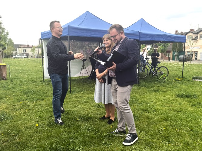 Zdjęcie przedstawia dwóch mężczyzn i jedną kobietę. Jeden z nich trzyma mikrofon i otwartą teczkę. W tle znajdują się dwa namioty wystawowe