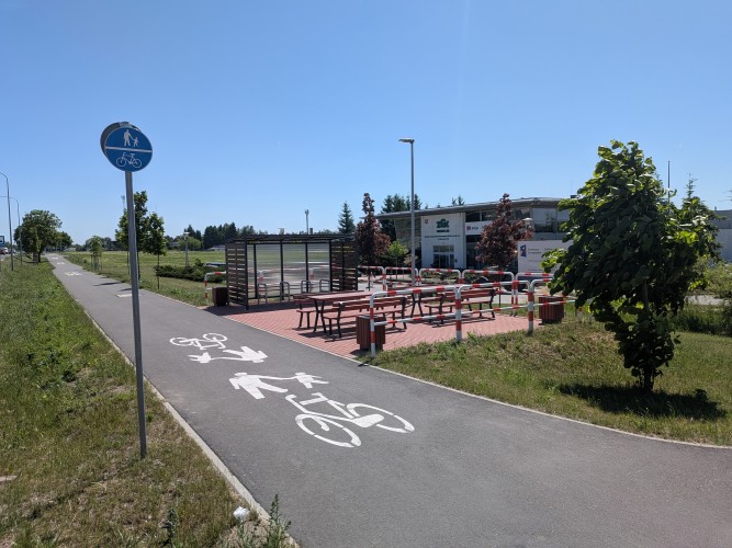 Zdjęcie przedstawiające ścieżkę rowerową oraz dla pieszych, wiatę rowerową oraz ławki ze stołami.