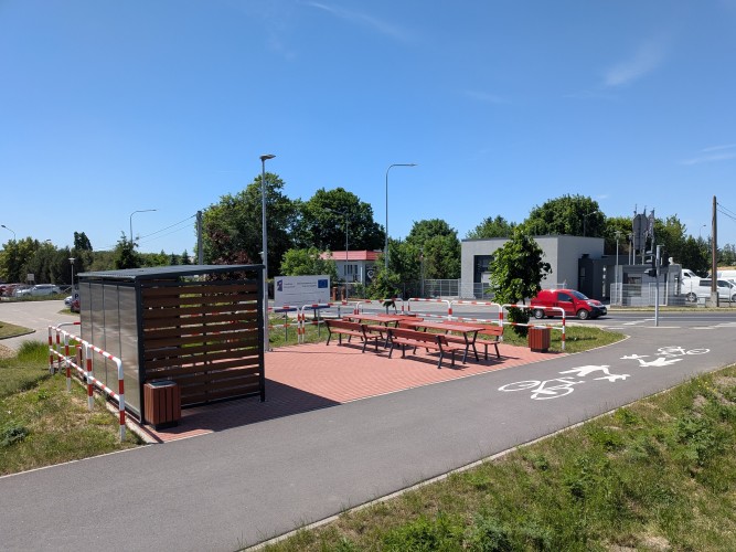 Zdjęcie przedstawiające ścieżkę rowerową oraz dla pieszych, wiatę dla rowerów oraz ławki ze stołami.