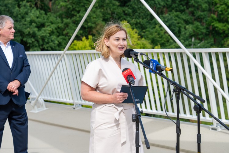 Kobieta stoi przed mikrofonami i udziela wywiadu. Za nią stoi mężczyzna.