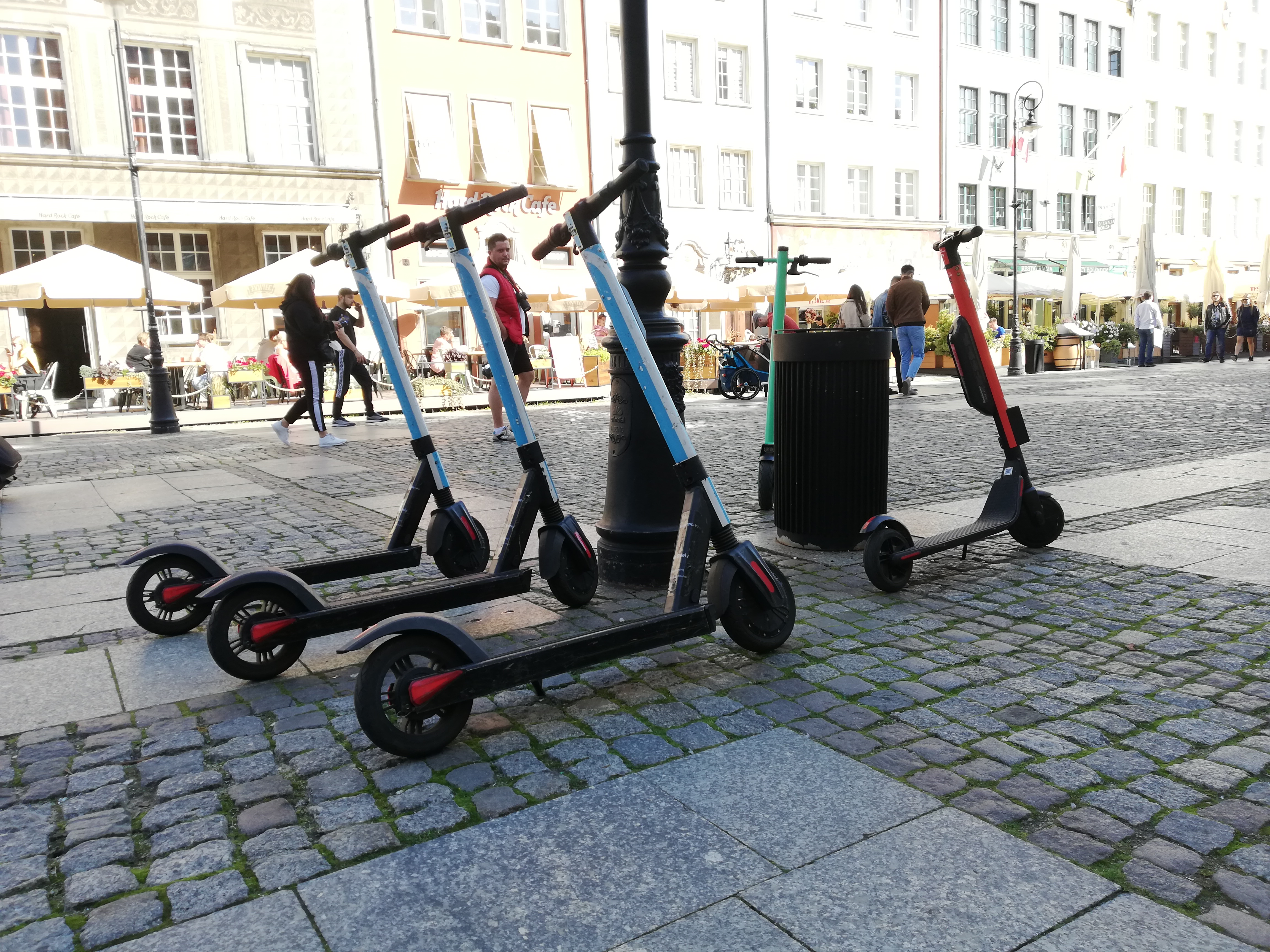 Zdjęcie przedstawia kilka elektrycznych hulajnóg miejskich zaparkowanych przy latarni i koszu na śmieci na brukowanej części starówki. W tle widoczne są kamienice, ogródki kawiarniane i przechodnie. Scena ilustruje mikromobilność jako element współczesnego transportu miejskiego.